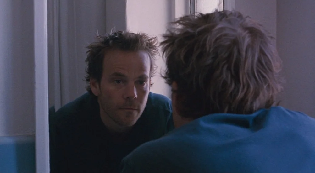 Somewhere (2010) movie still: medium — A man stands in front of a mirror in a small blue tiled room. He looks directly at his reflection in the glass.