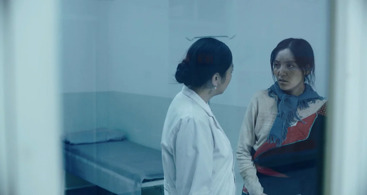 Balloon (2019) movie still: medium — A hospital room with a simple cot and white walls is viewed through glass. A female doctor in a white coat…