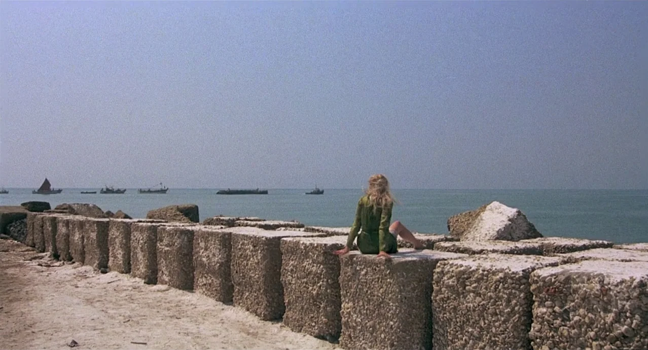 Amarcord (1973) movie still: wide — A seaside breakwater composed of large concrete cubes stretches into a calm, pale ocean. A lone woman sits on…