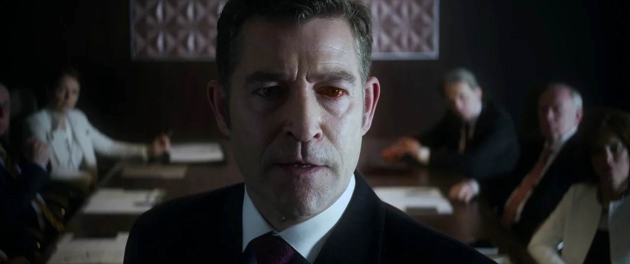 Mayhem (2017) movie still: closeup — A dark boardroom with a long wooden table surrounded by several figures in formal attire. The central man…