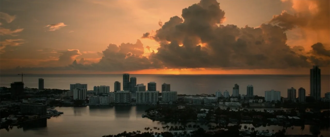 Bad Boys: Ride or Die (2024) movie still: establishing — A dense coastal city skyline sits along a dark calm bay at golden hour. Massive orange-lit…