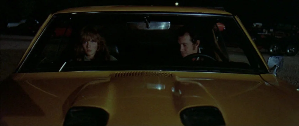 Two Lane Blacktop (1971) movie still: medium — A dark, empty space surrounds a yellow vehicle at night. The man drives while the woman looks forward…