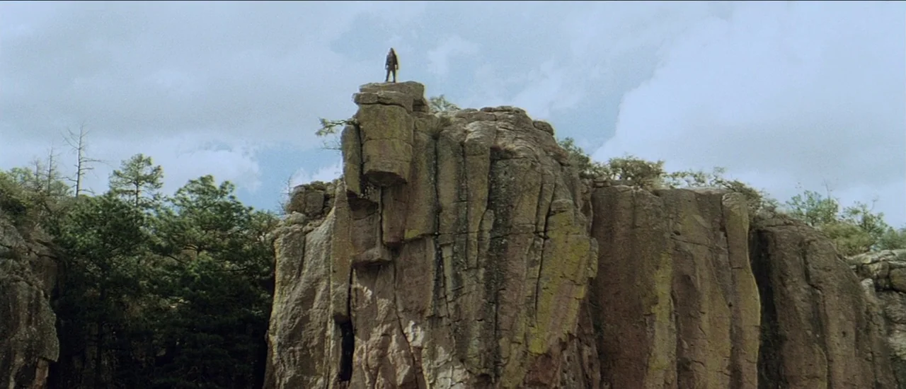 Renegade (2004) movie still: wide — A rugged cliff face rises against a cloudy sky with green trees at base. A lone person stands on the highest…
