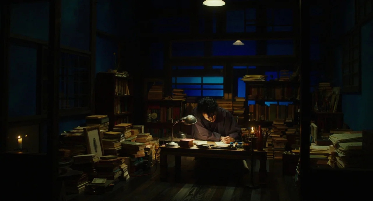 No Longer Human (2019) movie still: wide — A dark study filled with stacks of books on the floor and cluttered shelves. A man sits at a low table…