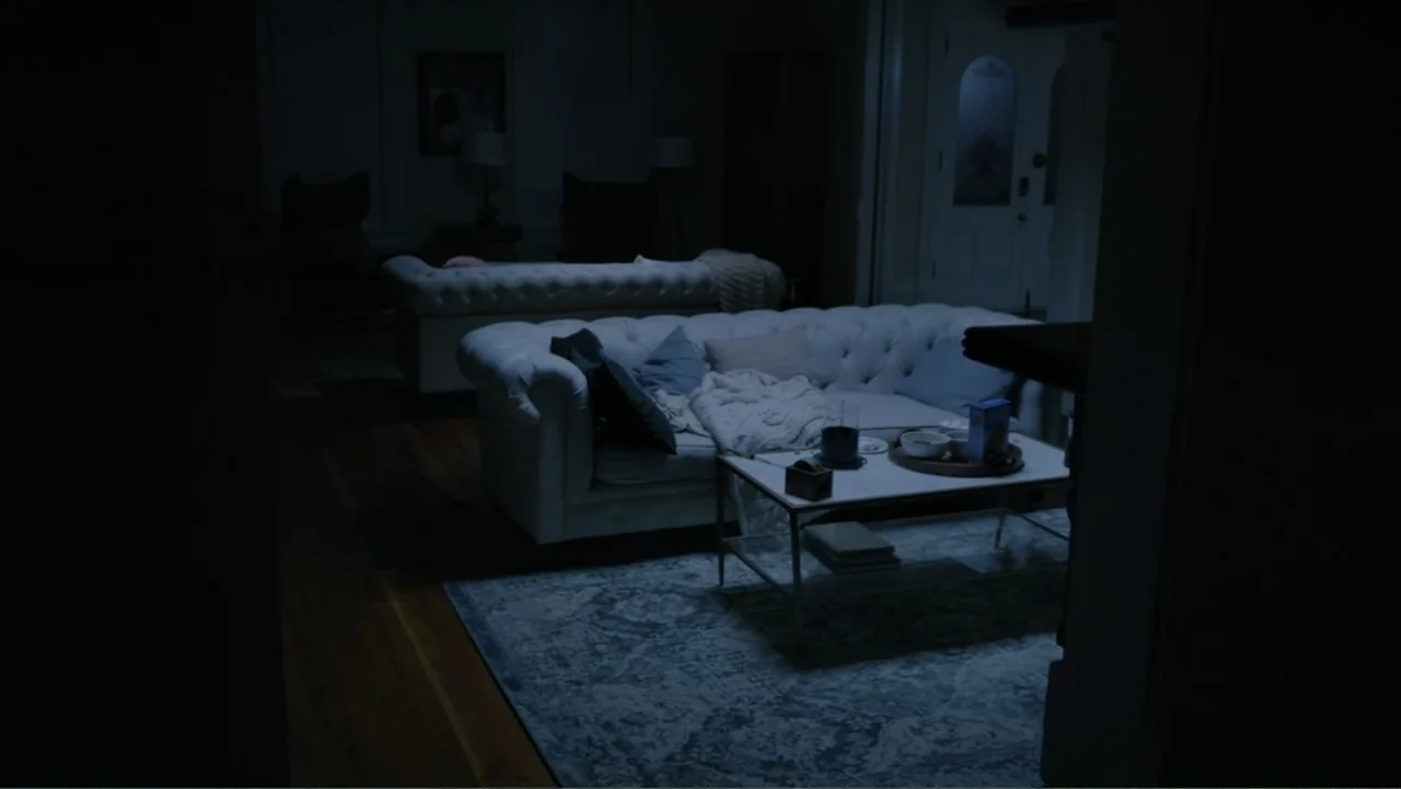 Storm Damage (2000) movie still: wide — A dark living room contains two white tufted sofas and a central coffee table. The space is sparsely lit…