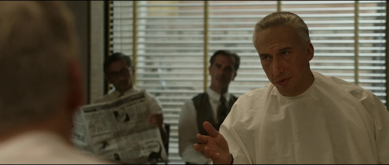 Ferrari (2023) movie still: medium — A 1940s setting with venetian blinds in the background and men seated in chairs. A man in a white cape talks…