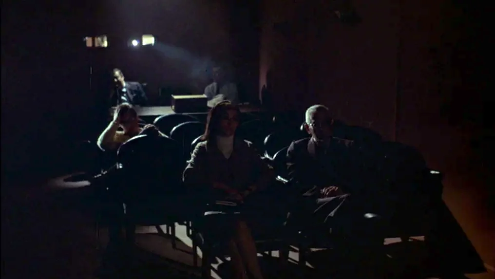 Targets (1968) movie still: medium — A dark, sparse room with rows of chairs facing a projector in the distance. Three people sit in the chairs…