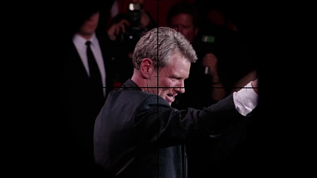 The Manchurian Candidate (2004) movie still: medium — A man in a dark suit stands amidst a crowd of photographers at night. He is waving his hand…