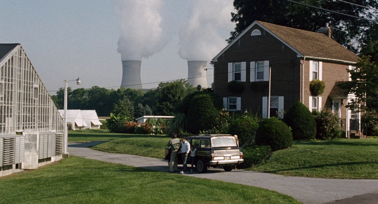 The Happening (2008) movie still: wide — A suburban brick house stands next to a driveway and a large glass greenhouse. In the distance two large…