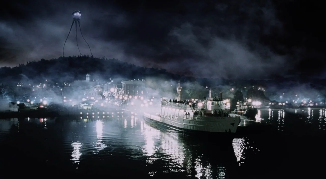 War of the Worlds (2005) movie still: establishing — A night scene shows a riverfront town illuminated by numerous scattered lights under a dark,…