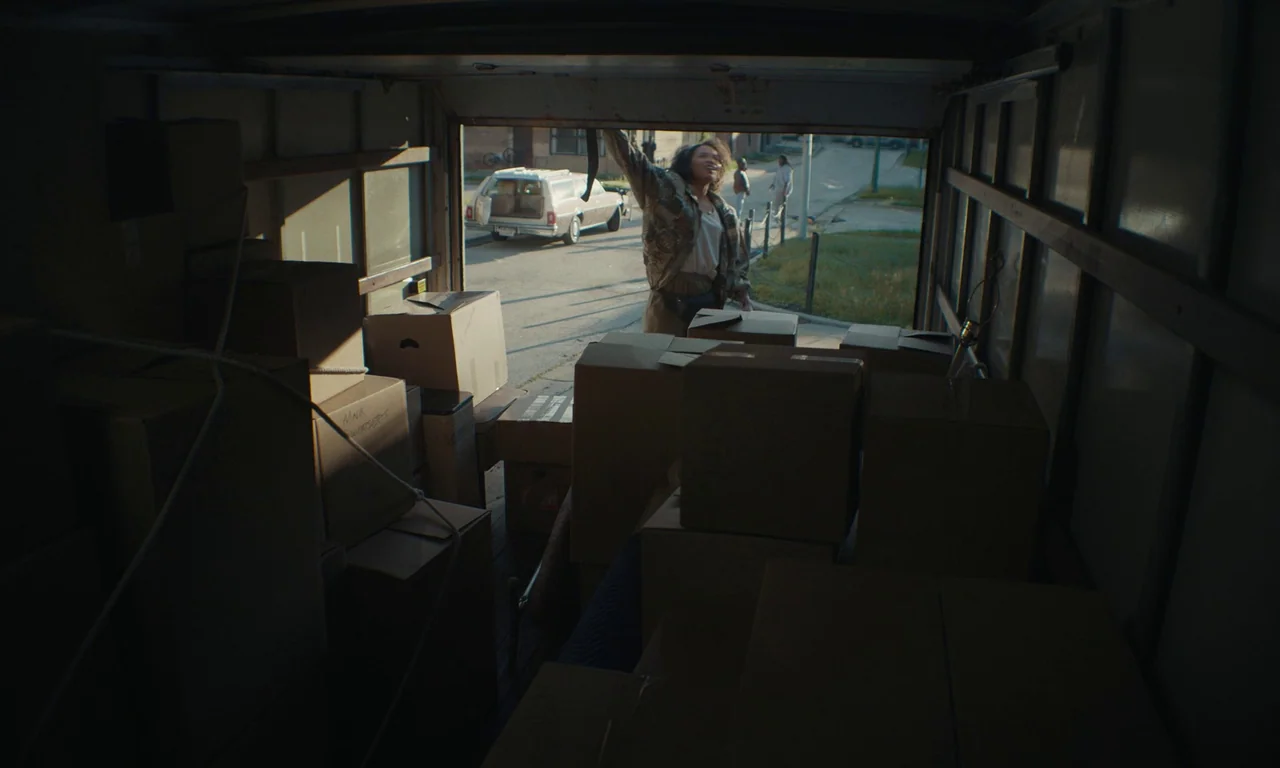 We Grown Now (2023) movie still: wide — The interior of a darkened rental truck is filled with cardboard packing boxes. A woman stands at the rear…