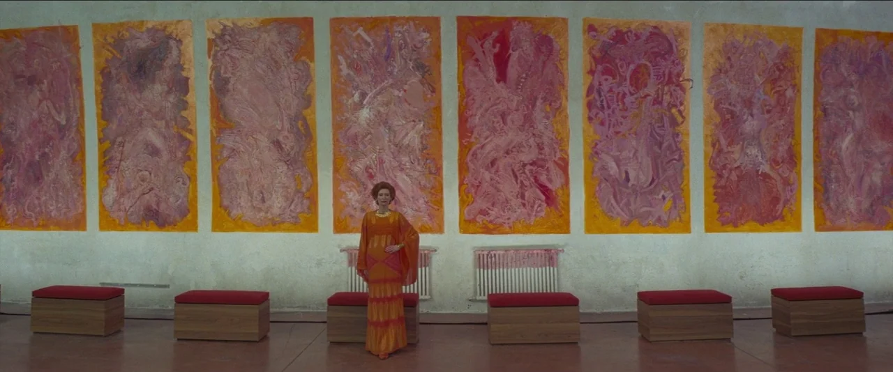 The French Dispatch (2021) movie still: fullbody — An art gallery wall is covered with six large vertical abstract paintings in orange tones. A…