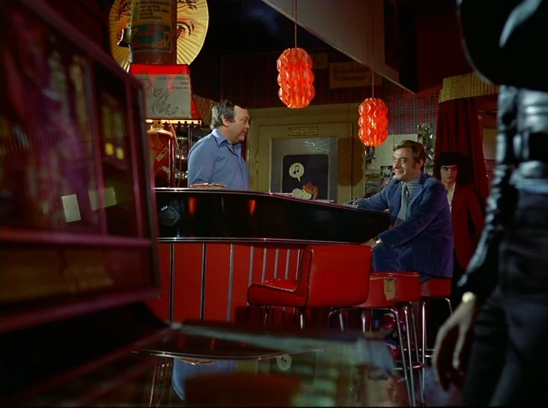 Fox and His Friends (1975) movie still: medium — A 1970s bar features red stools, red hanging spherical lamps, and wood-paneled walls. Two men…