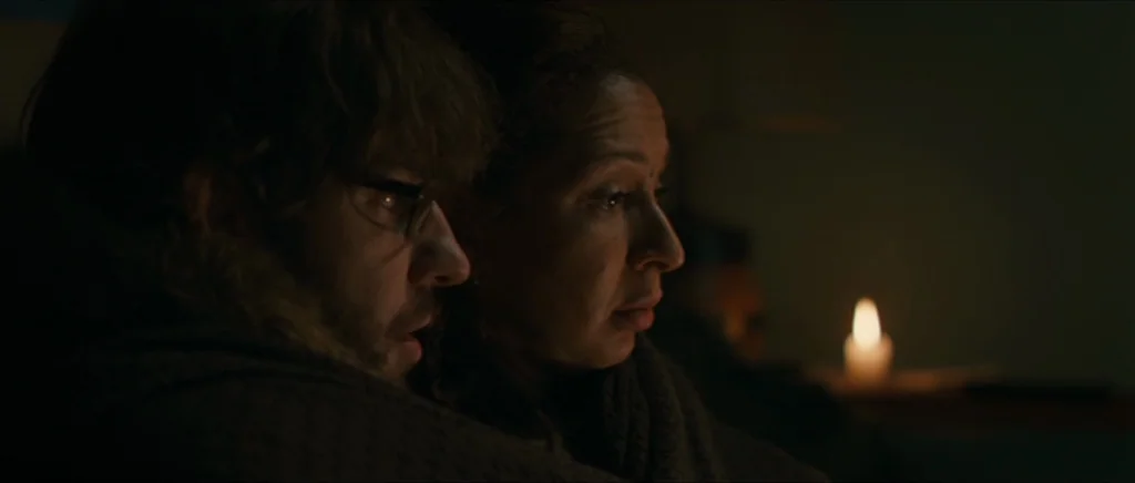 Away We Go (2009) movie still: medium — A dimly lit room at night with minimal illumination provided only by a candle. A man and a woman sit closely…