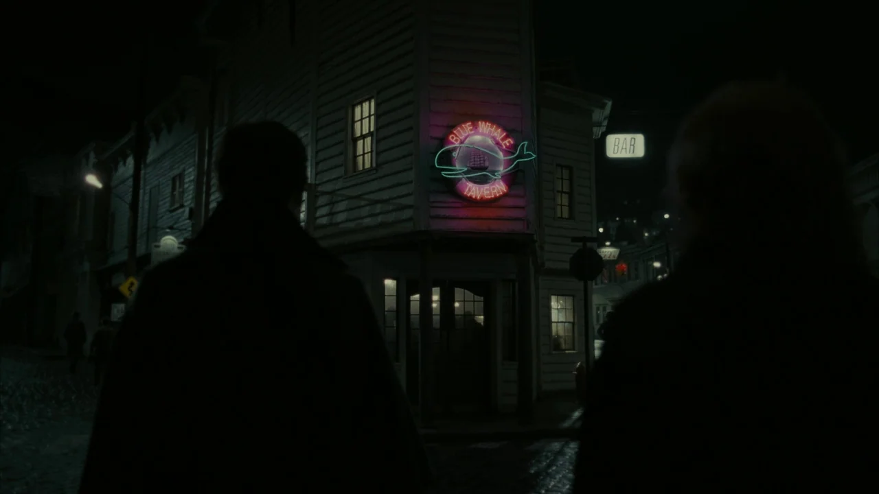 Dark Shadows (2012) movie still: wide — A dark, wet cobblestone street at night features a corner building with signage. Two silhouettes stand in…
