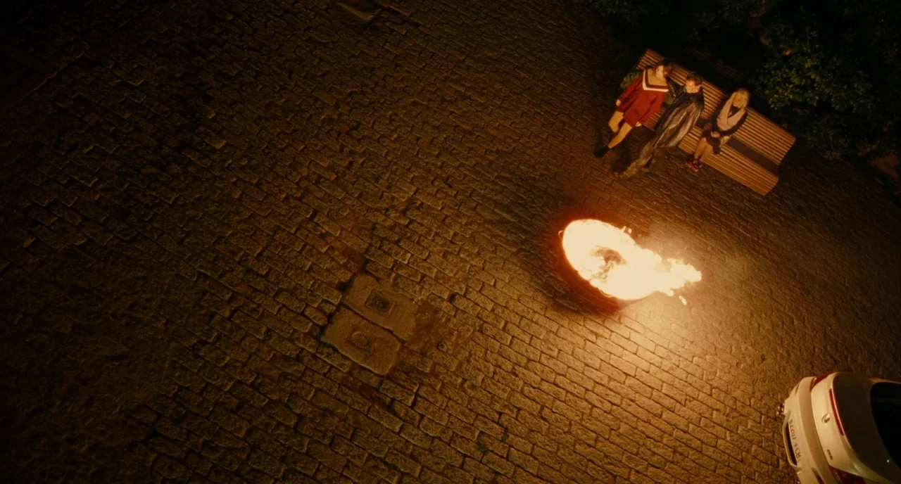 The Skin I Live In (2011) movie still: wide — A paved cobblestone area at night features a fire burning within a circular metal bin. Three…