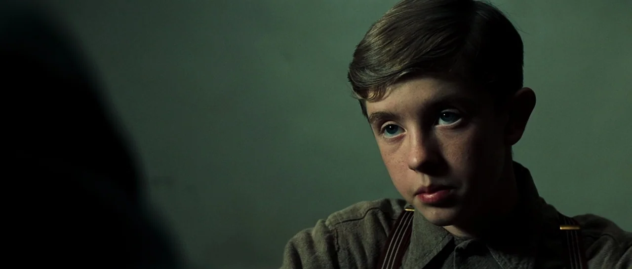 Changeling (2008) movie still: closeup — A dimly lit room with plain walls frames a child in a plain shirt. The boy looks toward someone off-screen…