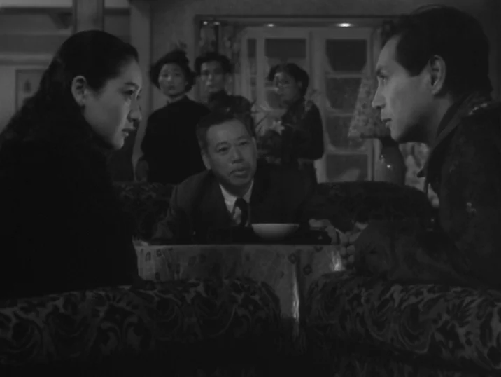 The Idiot (1951) movie still: medium — A dimly lit room with patterned furniture where three people sit around a table. An older man sits between…