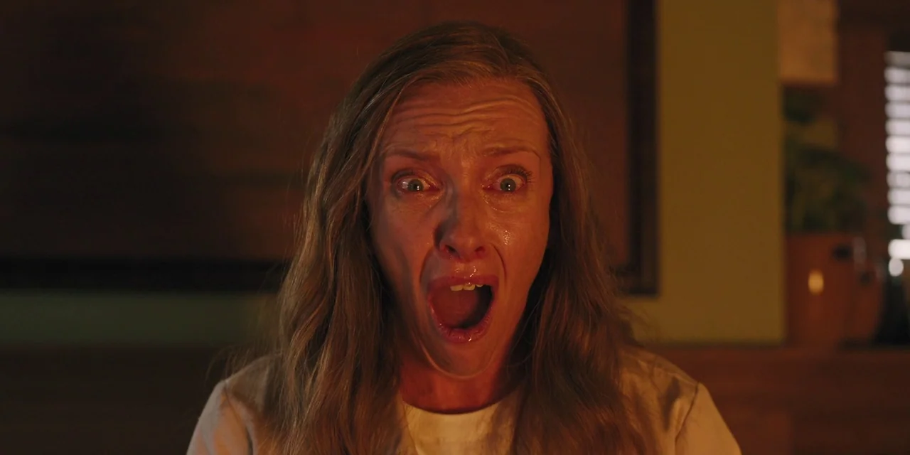 Hereditary (2018) movie still: closeup — A dark indoor setting features a woman in front of a blurred wall background. The woman stares forward with…