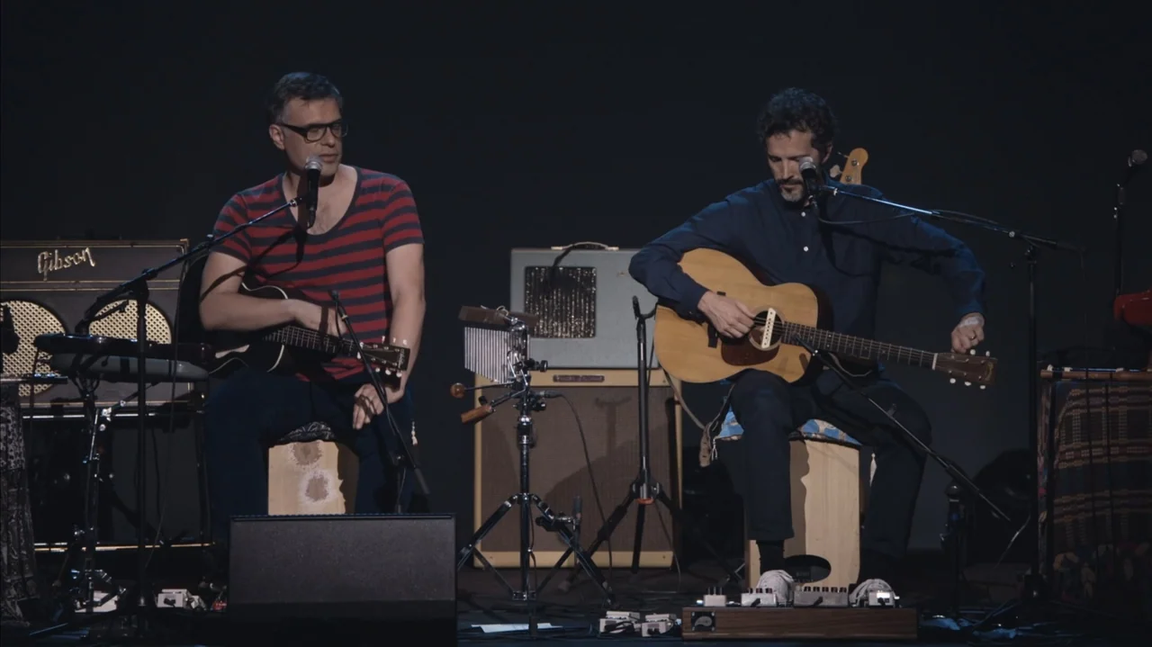Flight of the Conchords: Live in London (2018) movie still: medium — Two men sit on wooden boxes on a dark, sparsely decorated performance stage.…