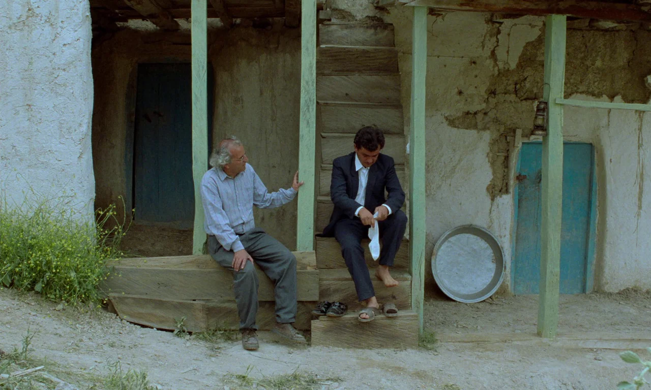 Through The Olive Trees (1994) movie still: fullbody — An exterior view of a rustic, mud-walled structure with light green painted wooden supports.…