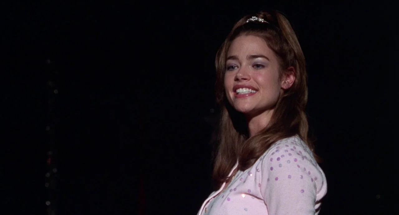 Drop Dead Gorgeous (1999) movie still: medium — A woman stands on a dark stage with a black backdrop behind her. She wears a sequined cardigan and…