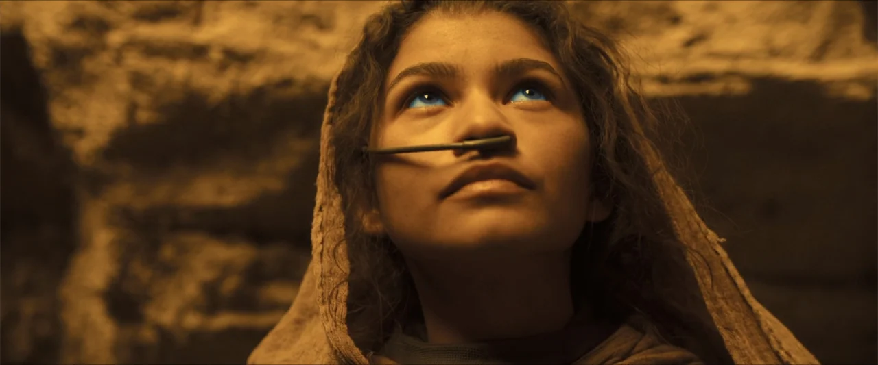 Dune (2021) movie still: closeup — A rocky underground chamber with coarse stone walls provides a barren, earthy environment. A young woman with…