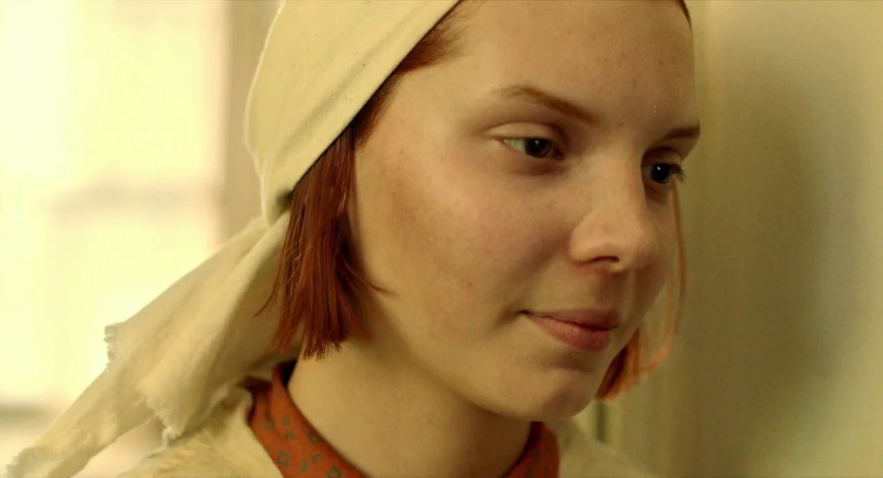 Beanpole (2019) movie still: closeup — A young woman stands inside a sparsely lit room wearing a simple cloth headcovering. She looks slightly to…