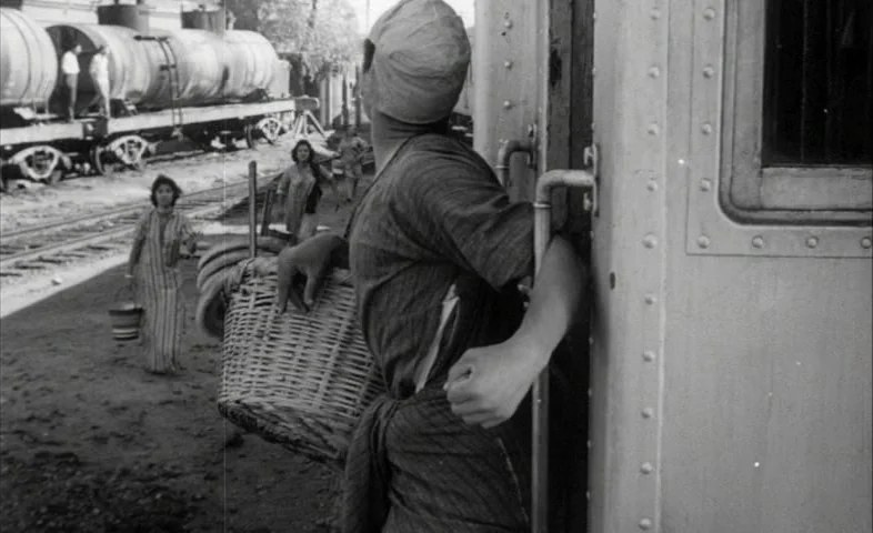 Cairo Station (1958) movie still: medium — A dusty railway track platform features industrial tanker cars in the background. A woman holds a large…