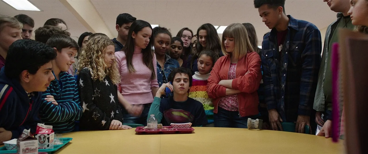 Shazam! (2019) movie still: medium — A group of diverse teenagers surrounds a central table in a school cafeteria setting. They observe a boy who is…