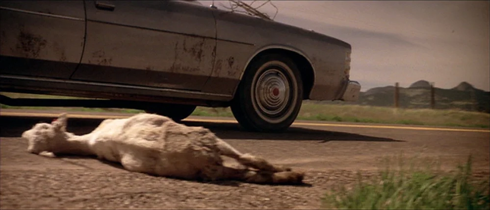 Close Encounters Of The Third Kind (1977) movie still: medium — A dusty, light-colored sedan sits on an asphalt highway in a remote, arid landscape.…