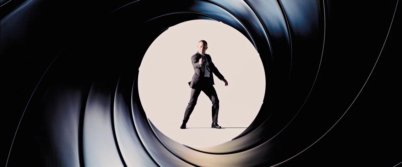 Skyfall (2012) movie still: fullbody — A man stands within a stylized, circular metal rifling pattern. The background is a flat, bright white surface.