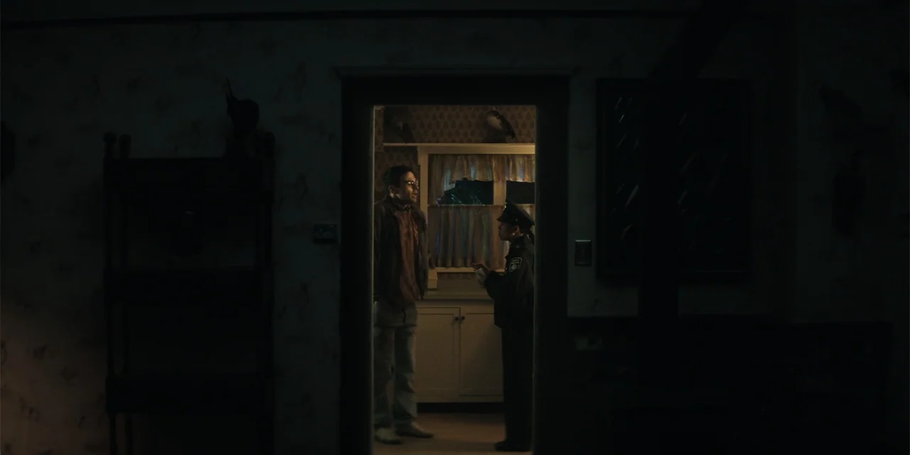 The Monkey (2025) movie still: medium — A dimly lit doorway connects a dark room to a brightly illuminated kitchen area. A man and a female police…