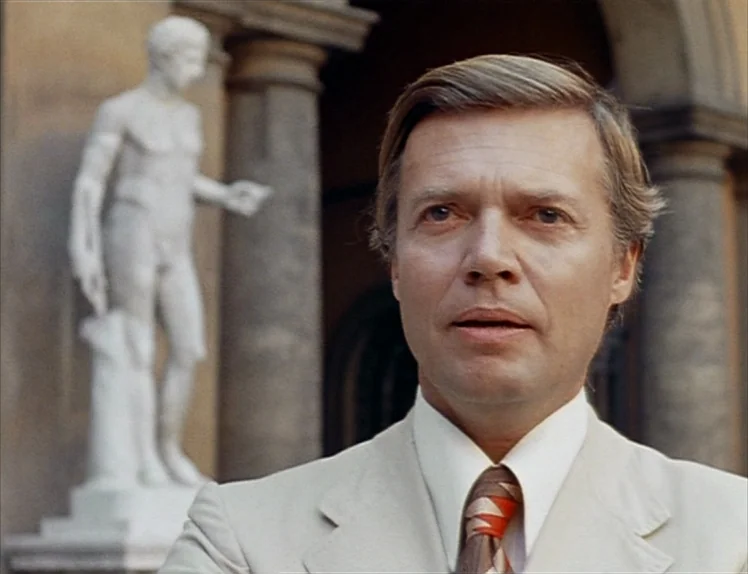 Martha (1974) movie still: closeup — An outdoor courtyard with classic stone architecture and a white marble statue in background. A middle-aged man…