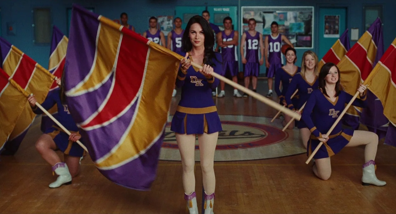 Jennifer’s Body (2009) movie still: fullbody — A group of cheerleaders in purple and gold uniforms stand on a wooden floor. The main subject holds a…