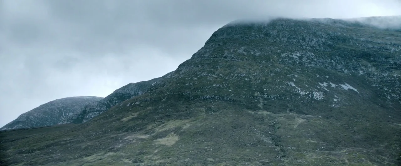 Black ’47 (2018) movie still: establishing — A rugged, rocky mountain slope rises against a heavy, overcast grey sky. Low clouds cling to the…