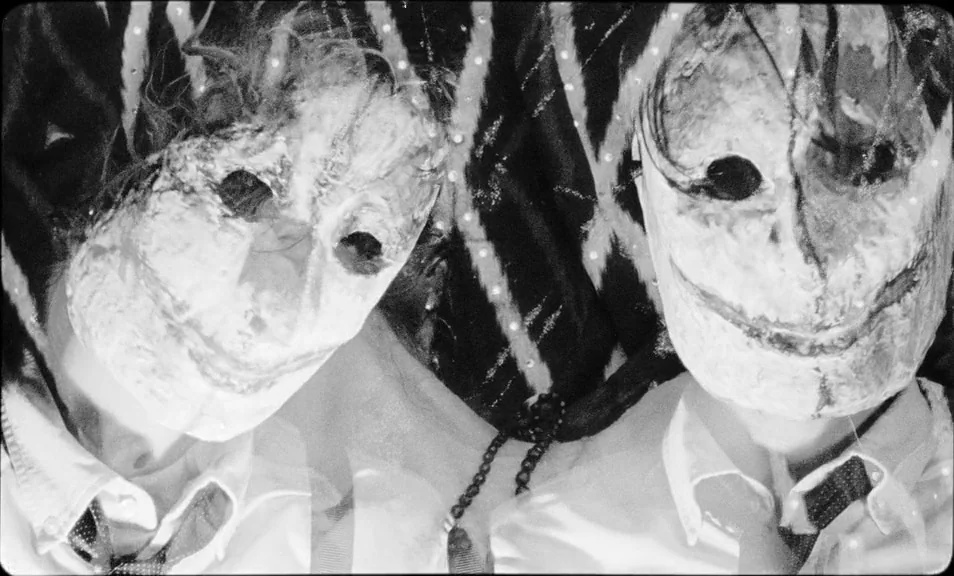 The Wild Boys (2017) movie still: closeup — Two individuals stand side by side wearing white textured masks in an indoor setting. The pair wear…