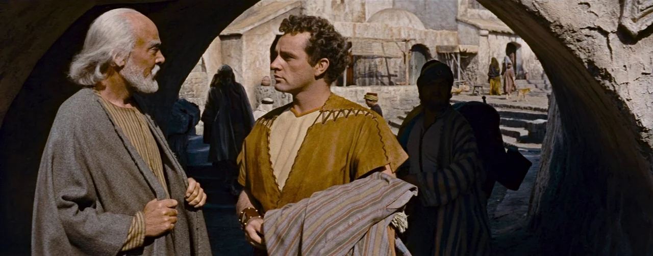 The Robe (1953) movie still: medium — A stone-walled courtyard is visible through a darkened archway in an ancient settlement. Two men stand beneath…
