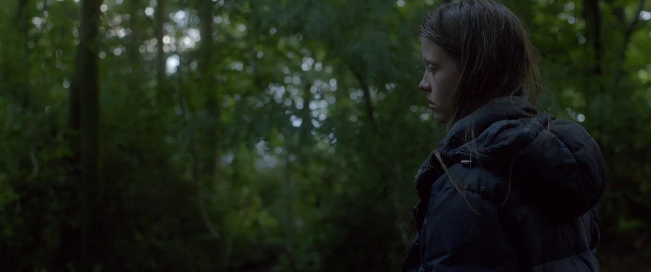 The Survivalist (2015) movie still: medium — A dense forest environment with thick green foliage and tall, dark tree trunks. A teenage girl wearing…