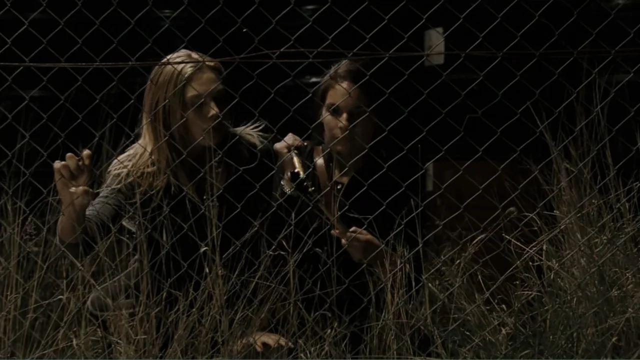 Tomorrow, When the War Began (2010) movie still: medium — Two women hide behind a metal chain link fence surrounded by tall dry grass. They are…