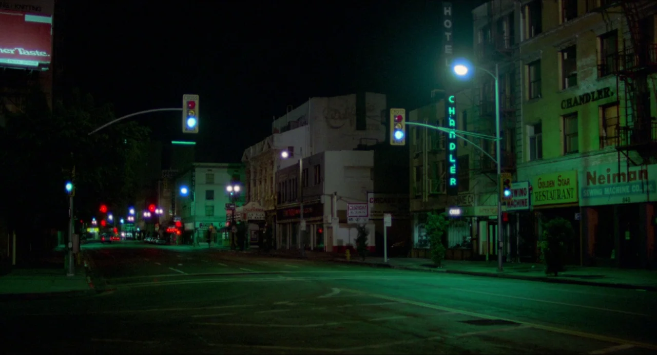 The Driver (1978) movie still: establishing — A quiet urban street intersection at night is lined with aged multi-story commercial buildings. Neon…
