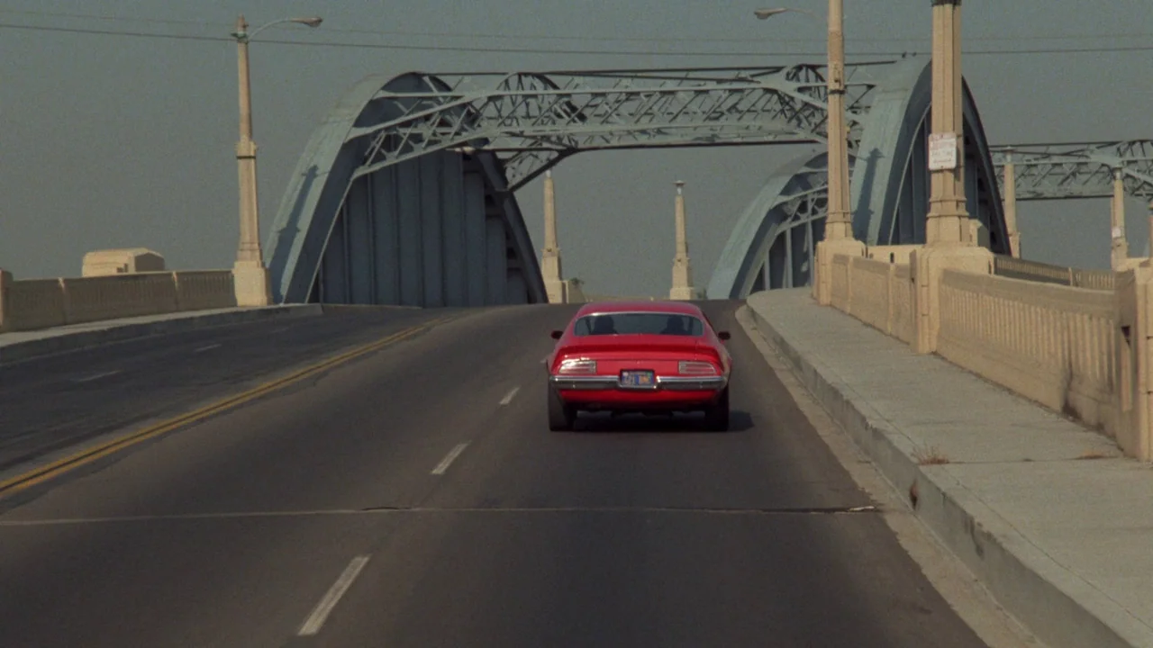 Repo Man (1984) movie still: wide — A long, arched steel bridge spans a road under a grey, overcast sky. A single red muscle car drives away from…