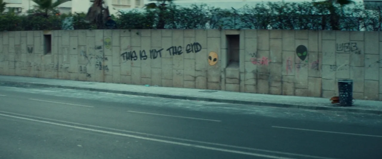 Animalia (2023) movie still: establishing — A long concrete wall lines a street, featuring various pieces of spray-painted graffiti. Alien faces and…