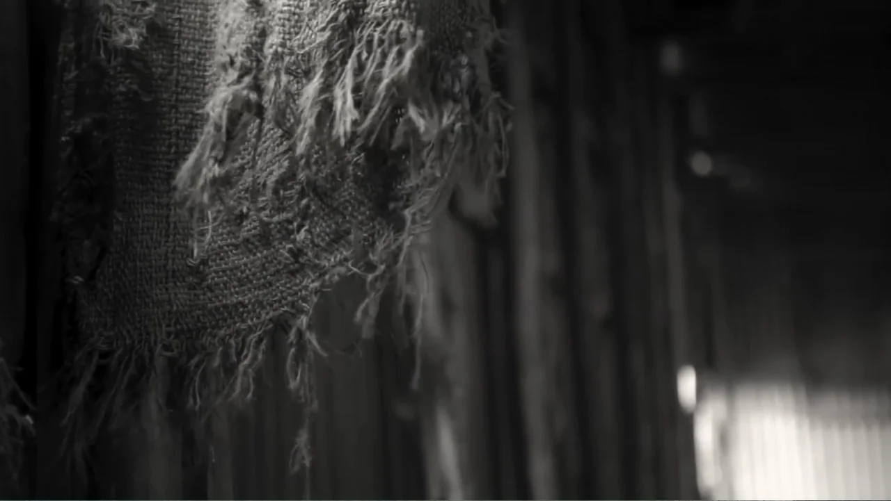 Faya Dayi (2021) movie still: closeup — A piece of frayed, coarse burlap fabric hangs from a weathered wooden wall. The fabric is out of focus in…