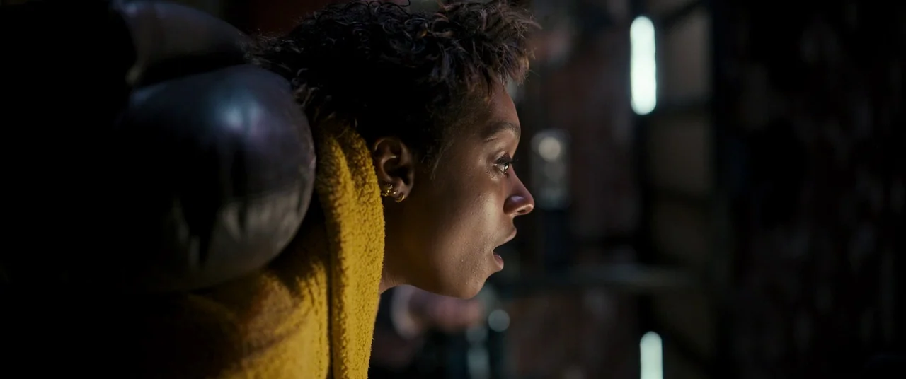 Talk To Me (2022) movie still: closeup — A dark, cramped interior with a leather headrest visible behind the subject. A woman stares into the…