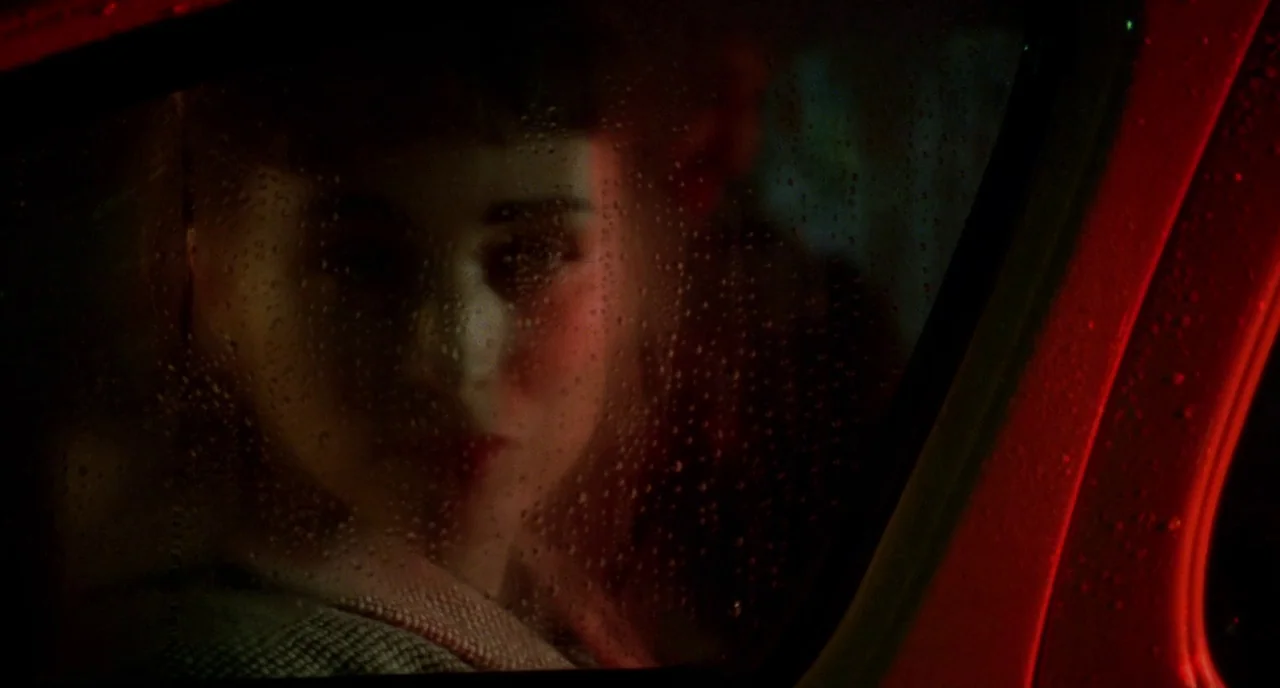 Carol (2015) movie still: closeup — A woman sits inside a car at night while rain beads on the window glass. The interior is dark with a vivid red…