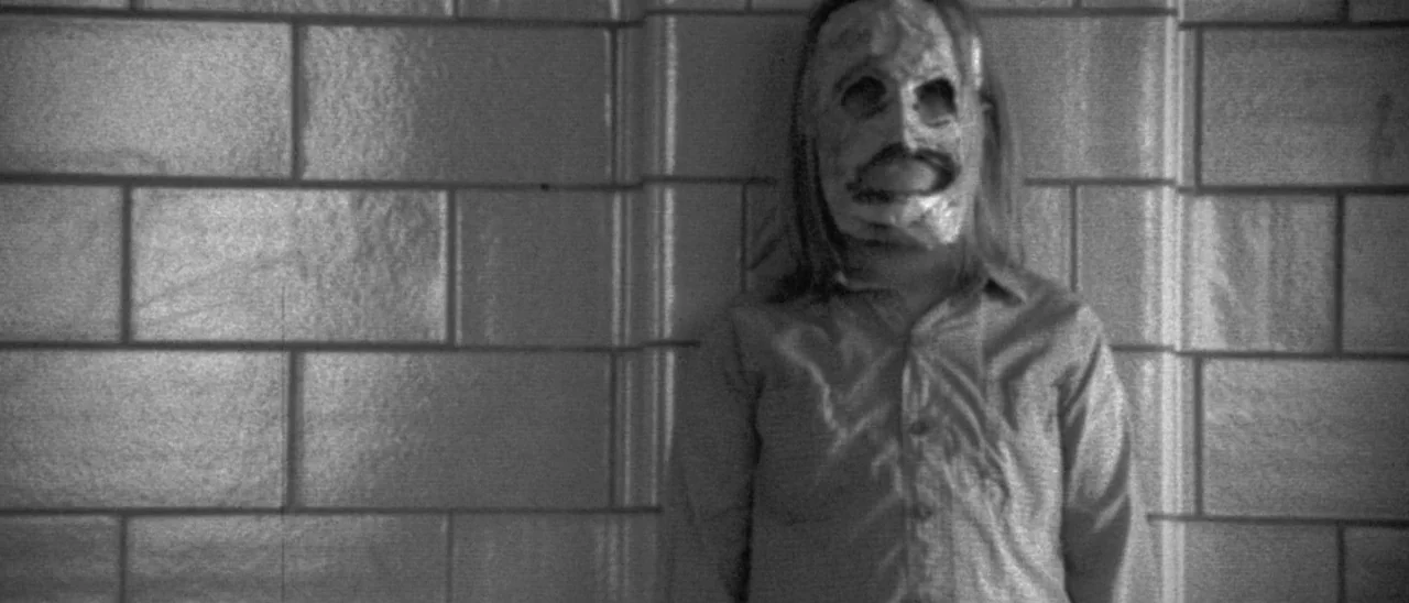 Halloween (2007) movie still: medium — A person in a mask stands against a tiled wall in a dimly lit hallway. The individual remains motionless,…