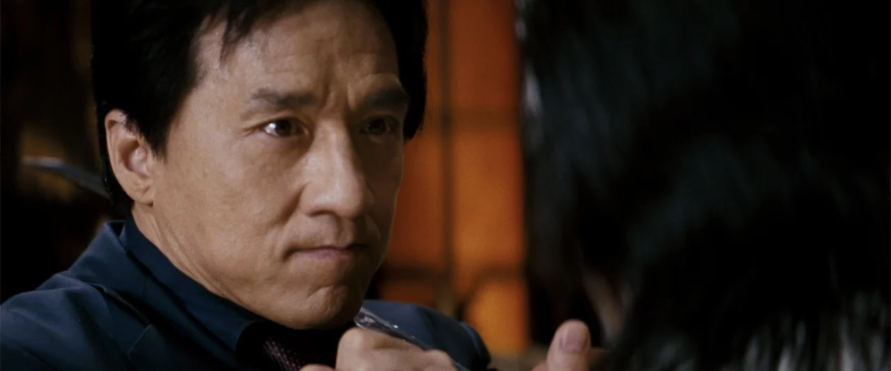 Rush Hour 3 (2007) movie still: closeup — A dimly lit indoor space with blurred background elements suggesting a seated dining area. An older East…