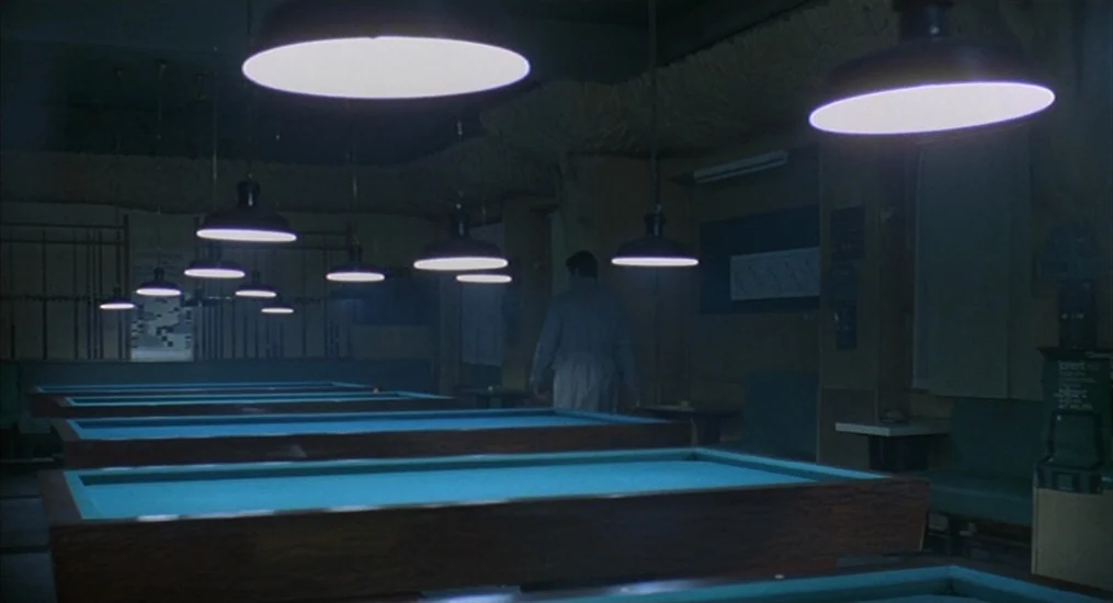 Le Cercle Rouge (1970) movie still: wide — A dimly lit billiards hall features several tables with bright blue felt surfaces under pendant lights. A…