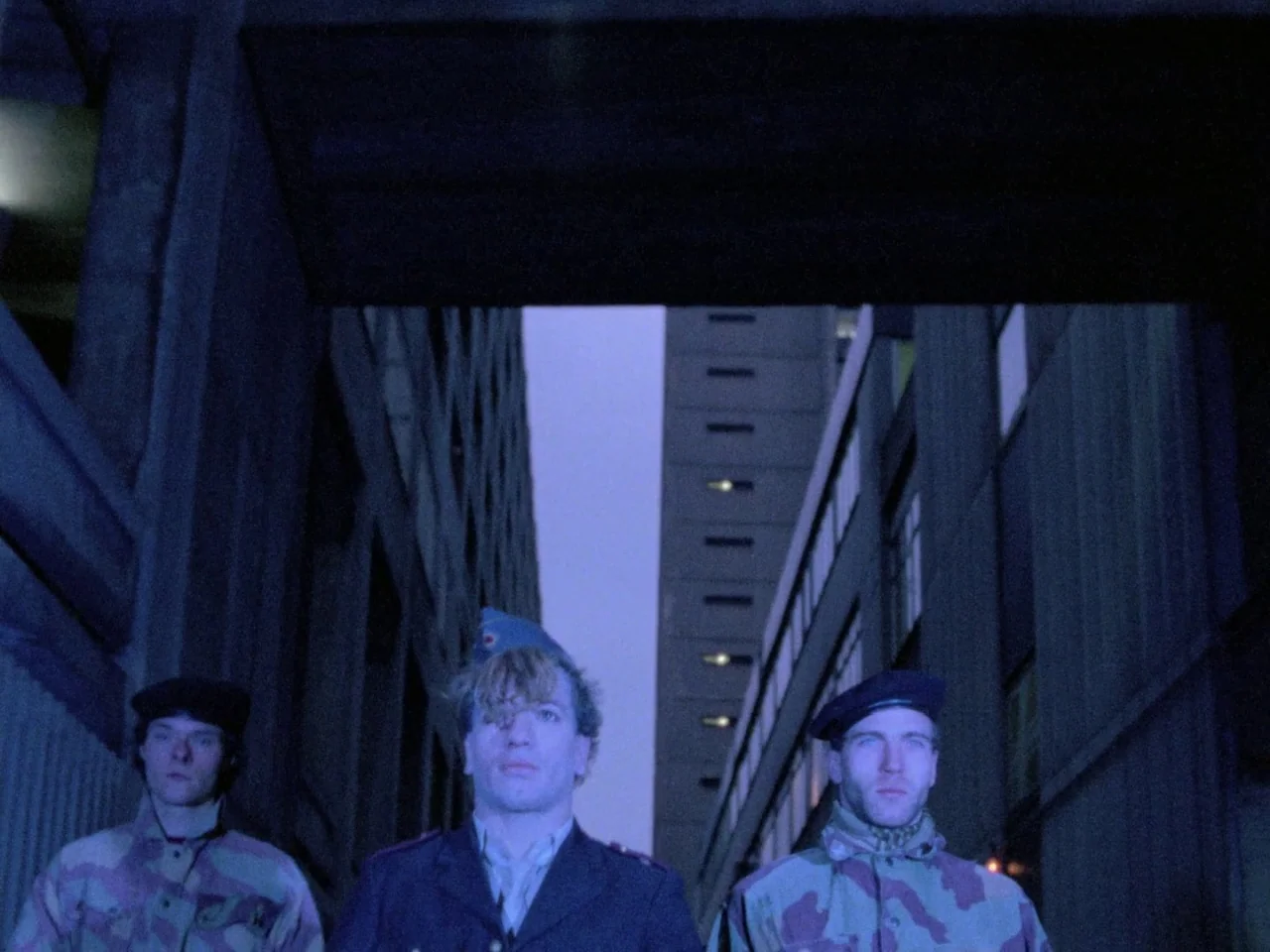 Decoder (1984) movie still: medium — A dark urban alleyway is framed by tall, imposing concrete buildings rising into the night. Three men dressed…